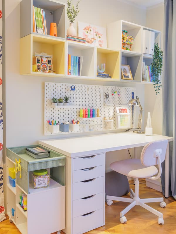 A well-organised children's study area featuring a white desk with drawers, a swivel chair, pegboards and open shelving filled with books, stationery and decorative items.