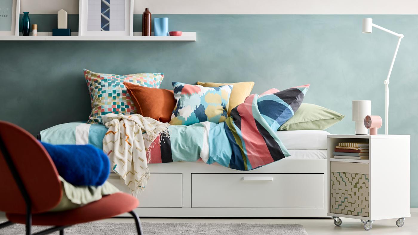 A well-organized bedroom scene with sleep storage solutions, neutral bedding, and a mic of colourful cushions. Includes a mirror, and a small table lamp adding functional accents to the space.