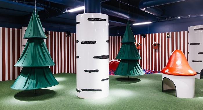 A whimsical indoor children's play area designed like a forest, featuring soft artificial trees, a large white tree trunk with black markings, a red mushroom-shaped structure, and a ball pit in the background. The walls are decorated with red and white vertical stripes.