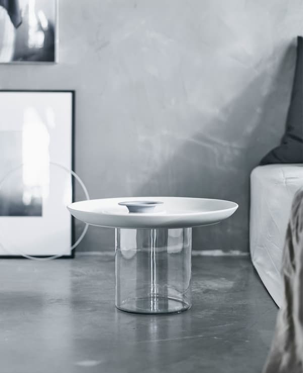 A white and clear bedside table next to a bed in a grey room.