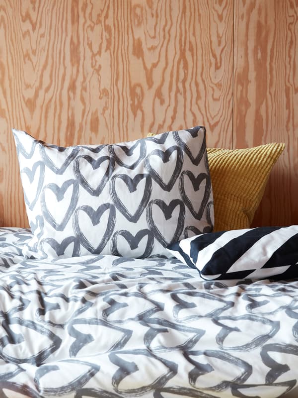 A white and gray LYKTFIBBLA quilt with a heart pattern on lies on a bed in a wood-panelled modern bedroom.