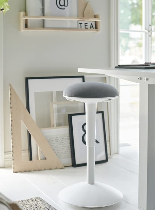 A white and grey stool next to a tall white desk, with artwork leaning against the wall behind.