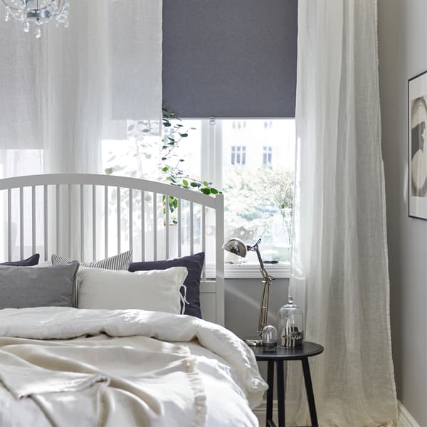A white bed frame, white curtains, a gray block-out roller blind, a chandelier and a black nest of tables.