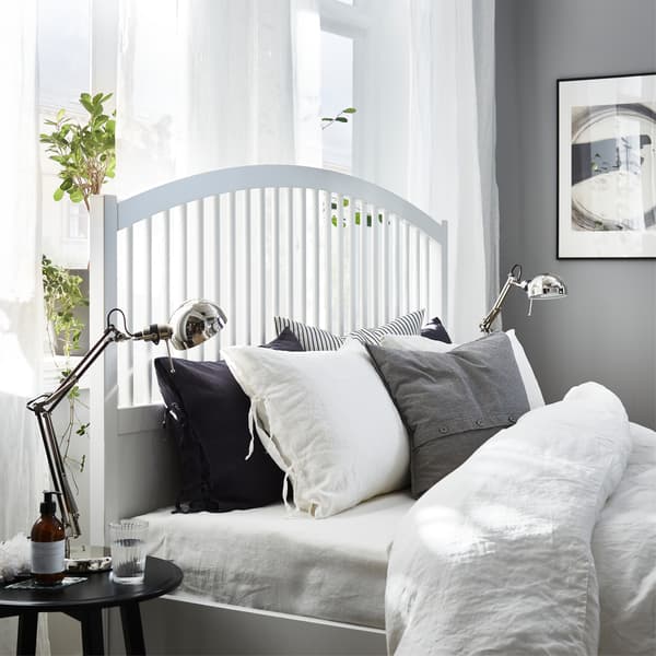 A white bed frame, white curtains, white, grey and black bed textiles and nickel-plated work lamps.