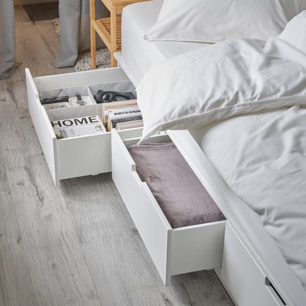 A white bed frame with two spacious, open drawers at the bottom. A pink throw, books, glasses and a watch are stored inside.