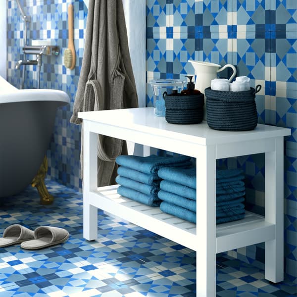A white bench with towels on a lower shelf and accessories on top, with blue tiled walls and floor.