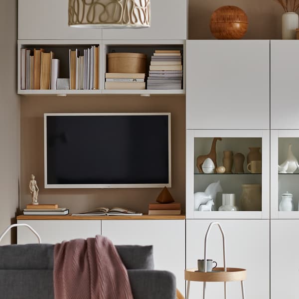 A white BESTÅ TV bench is beneath a wall-mounted TV, with other BESTÅ storage system units above and on one side.