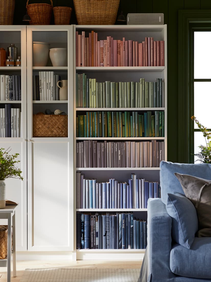 A white BILLY bookshelf filled with books arranged by color in a rainbow gradient, with decorative baskets and vases on the left and a blue sofa partially visible on the right.