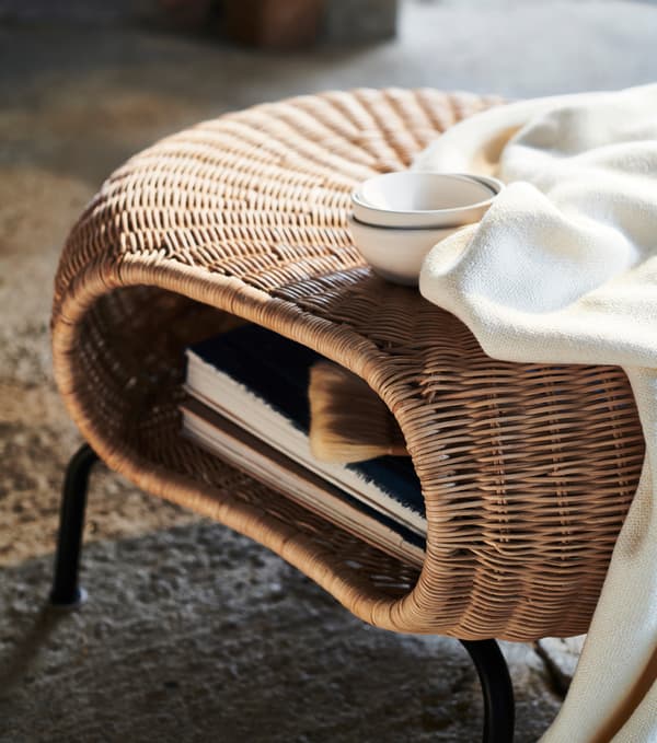 A white blanket and a cup sit on top of a rattan footstool that contains several magazines