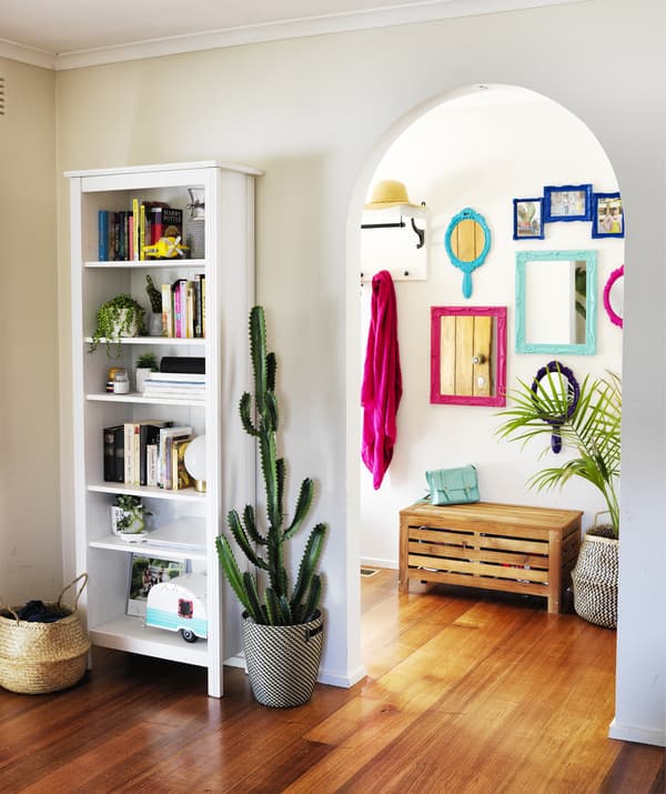A white bookcase and large cactus next to an archway leading through to a hallway with colourful mirrors on the wall.