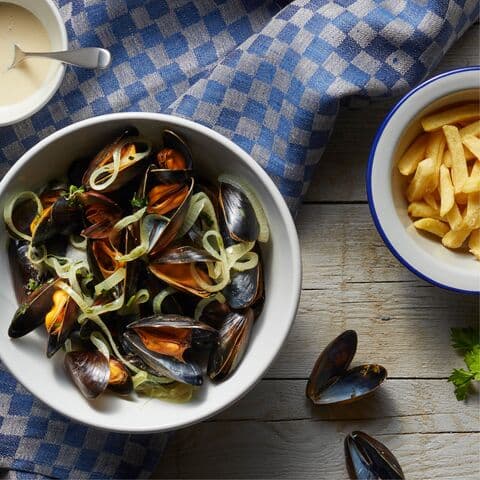 A white bowl with mussels, a medium white bowl with fries and a small white bowl with mussel sauce. Underneath the bowls is a blue kitchen towel with checkered pattern.