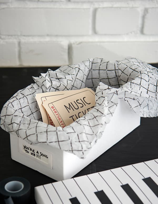 A white box is decorated like a piano keyboard and filled with decorative tissue paper and two tickets to a concert.