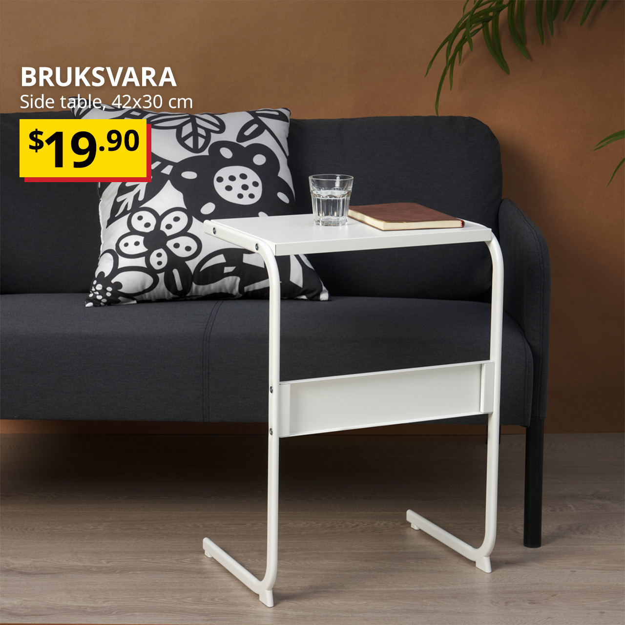 A white BRUKSVARA side table in white placed infront of a black GLOSTAD sofa in a living room setting. Words on image reads "BRUKSVARA, Side table, 42x30 cm, $19.90" 