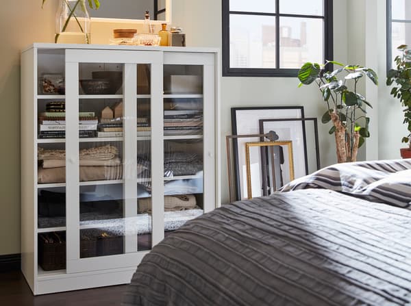 A white cabinet with sliding glass doors, frames that stand against a wall and a gray bedspread.