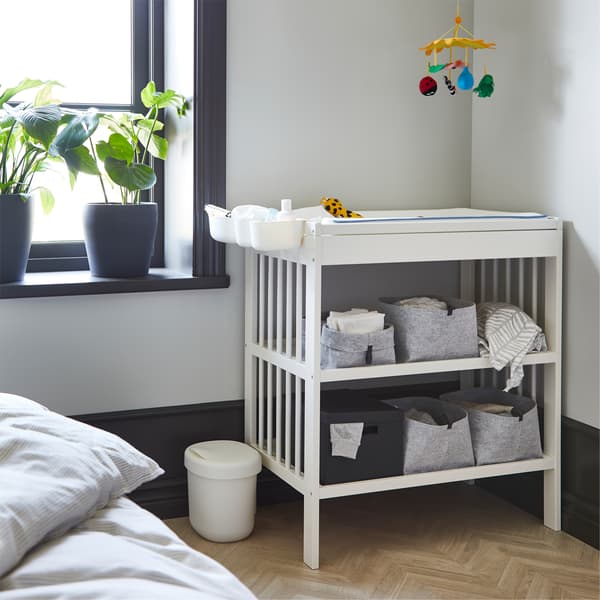 A white changing table with grey open boxes on its shelves. Beside is a window with dark grey plant pots on the window sill.