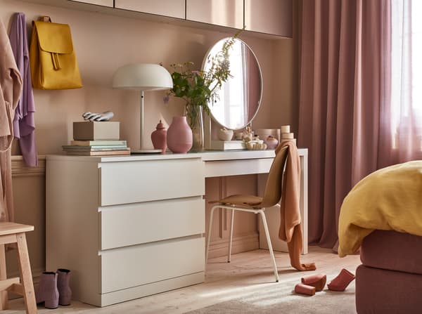 A white chest of drawers, a white dressing table, a white table lamp, pink small vases and a white round table mirror.
