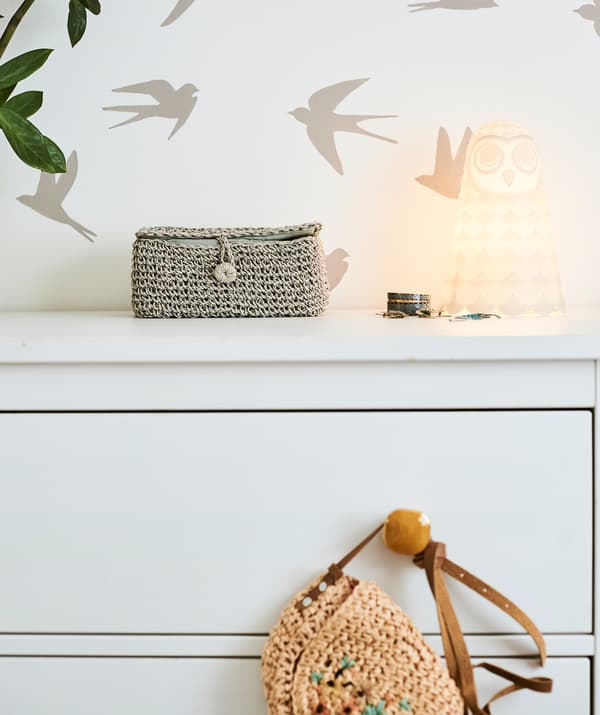 A white chest of drawers with amber-colored handles and an owl light on top, pushed against a wall with birds stencil motif.