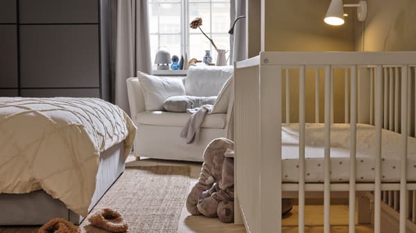 A white cot opposite the end of a grey bed, with a white BACKSÄLEN 1.5 seat armchair behind it near a window.