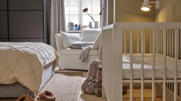 A white cot opposite the end of a grey bed, with a white BACKSÄLEN 1.5 seat armchair behind it near a window.