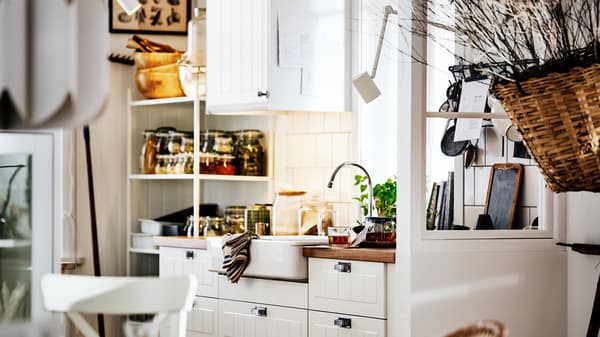 A white country-style kitchen with STENSUND fronts on METOD cabinets, a white sink and shelving with jars and utensils.