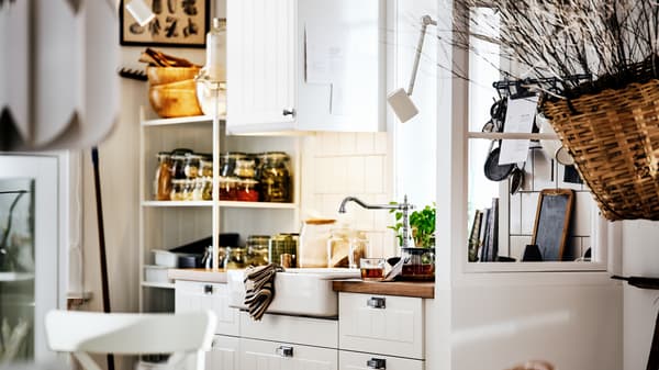 A white country-style kitchen with STENSUND fronts on METOD cabinets, a white sink and shelving with jars and utensils.