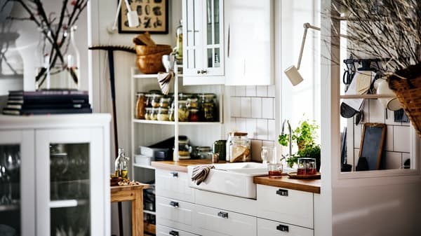 A white country-style kitchen with STENSUND fronts on METOD cabinets, a white sink and shelving with jars and utensils.