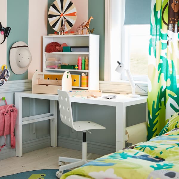 A white desk, a white children's desk chair, a white table lamp and curtains in a jungle pattern.