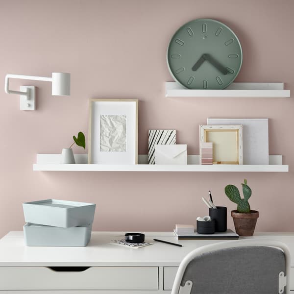 A white desk against a pink wall, with pictures and a clock displayed on white picture ledges.