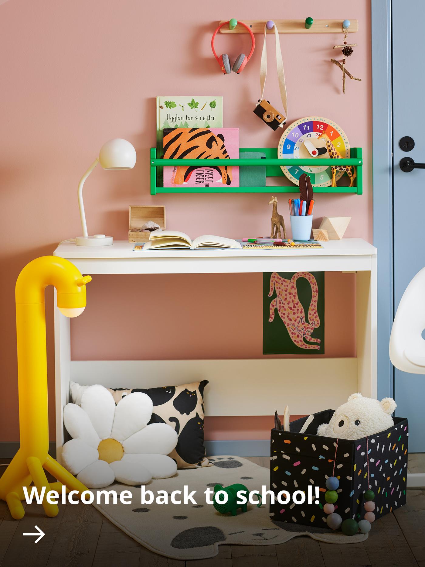 A white desk and chair in a children’s room, featuring a giraffe-shaped yellow floor lamp, a flower-shaped cushion, a patterned rug and a black storage box filled with toys.
