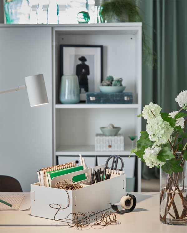 A white desk organiser is placed on a white desk in a bedroom, and it holds pens, scissors, notebooks and more.