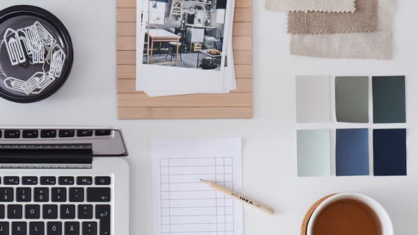 A white desk with a laptop, two cups of tea, a checklist, a wooden IKEA pencil and colour swatches.