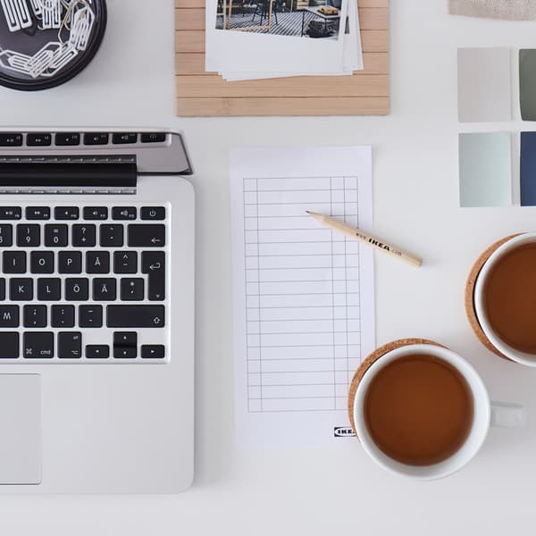 A white desk with a laptop, two cups of tea, a checklist, a wooden IKEA pencil and color swatches.