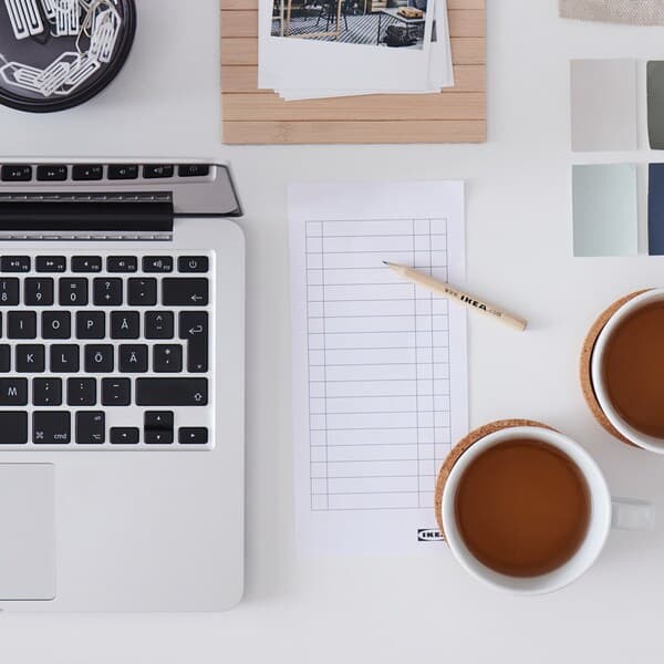 A white desk with a laptop, two cups of tea, a checklist, a wooden IKEA pencil and color swatches.