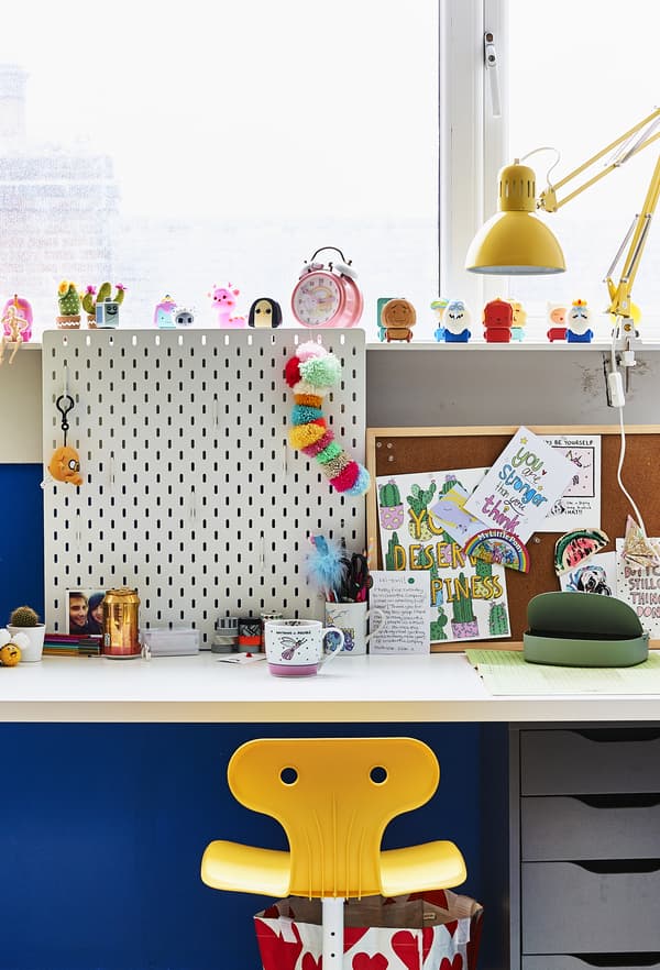 A white desk with a white pegboard on top shown together with yellow swivel chair and work lamp.