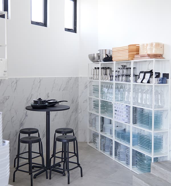 A white display shelf with various plates, bowls and other kitchen gadget. A black side table with four black chairs are placed against a grey wall.