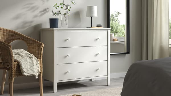 A white dresser in a bedroom with a plant sitting on top and a wicker chair next to it.