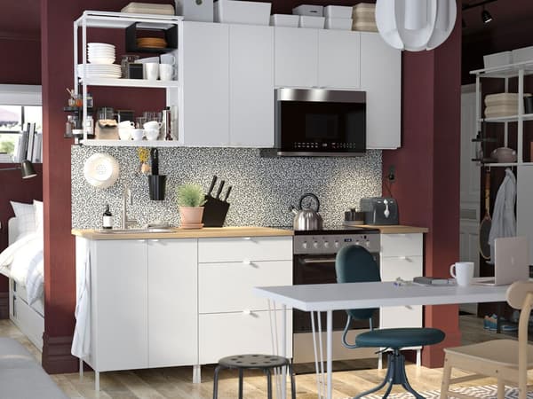 A white ENHET kitchen combination along a wall in a studio apartment with a desk in front and a bed behind the wall.