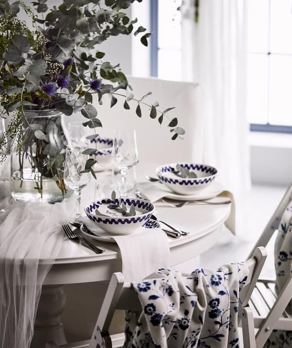 A white, festive round table with a bouquet of wild flowers in a glass vase, and white and blue bowls and plates.