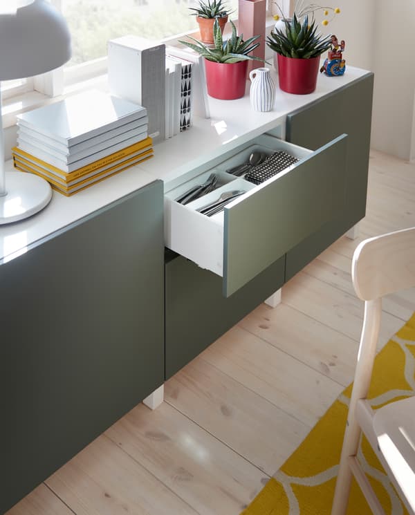A white/grey-green storage combination with plants, books and a table lamp on its top surface. Cutlery is stored in a drawer.