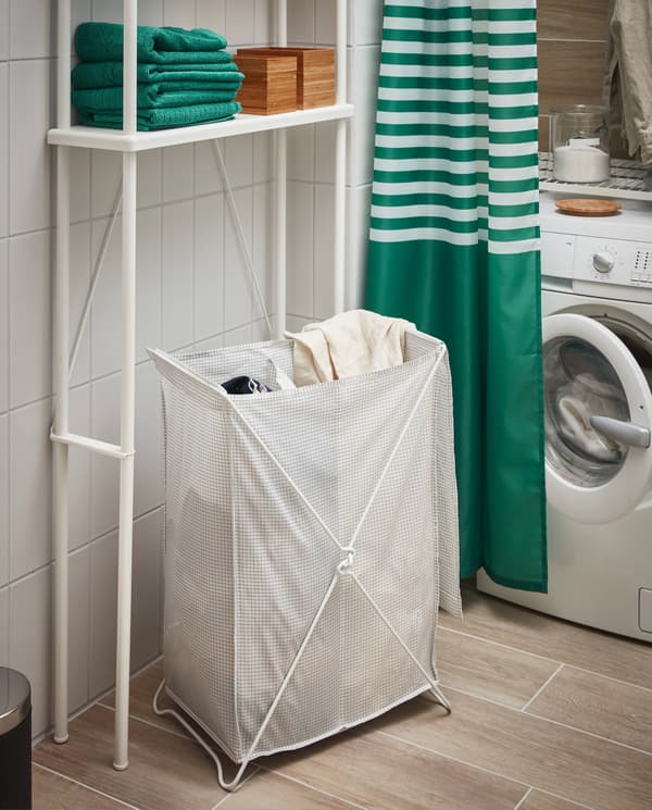 A white/grey laundry basket, a laundry machine, a green/white shower curtain, green towels and DYNAN open storage in white.