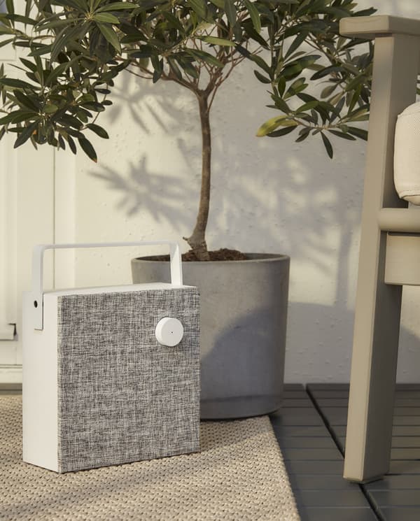 A white/grey, square and cordless speaker with handle stands on a beige rug next to a grey outdoor armchair.