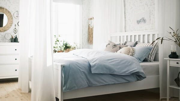 A white HEMNES bed in a bright room surrounded by sheer white SILVERLÖNN curtains, with cushions and blue bed linen on it.
