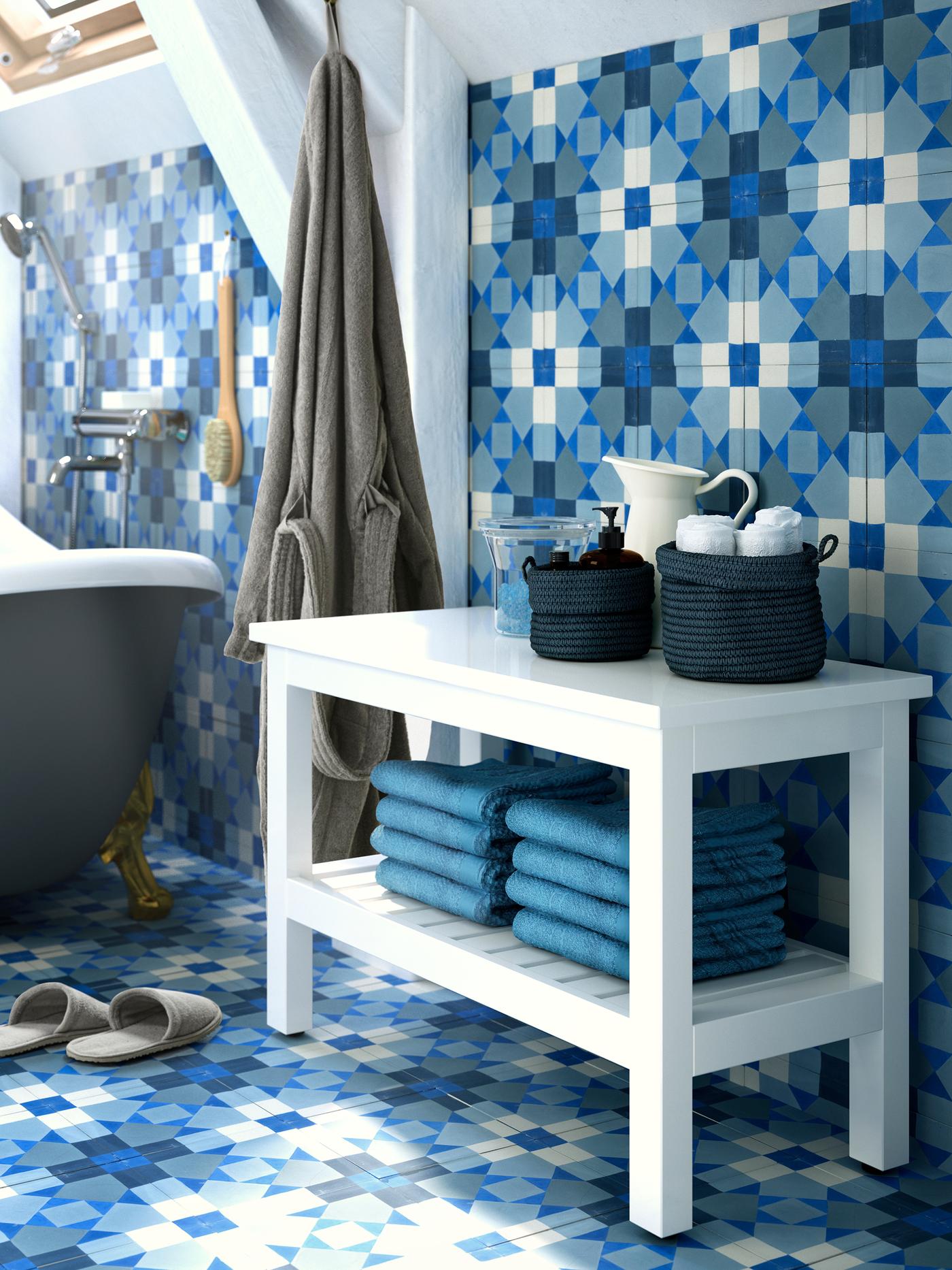 A white HEMNES bench with towels on a lower shelf and baskets with accessories on top, with blue tiled walls and floor.
