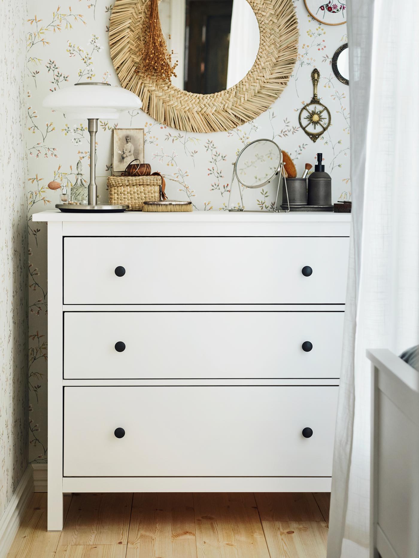 A white HEMNES chest with 3 drawers with a white table lamp on it, in front of a floral wall with a VRIGSTAD mirror on it.