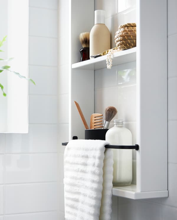 A white HEMNES wall shelf holding bottles, makeup and a white towel on a black metal rack attached to the shelf.