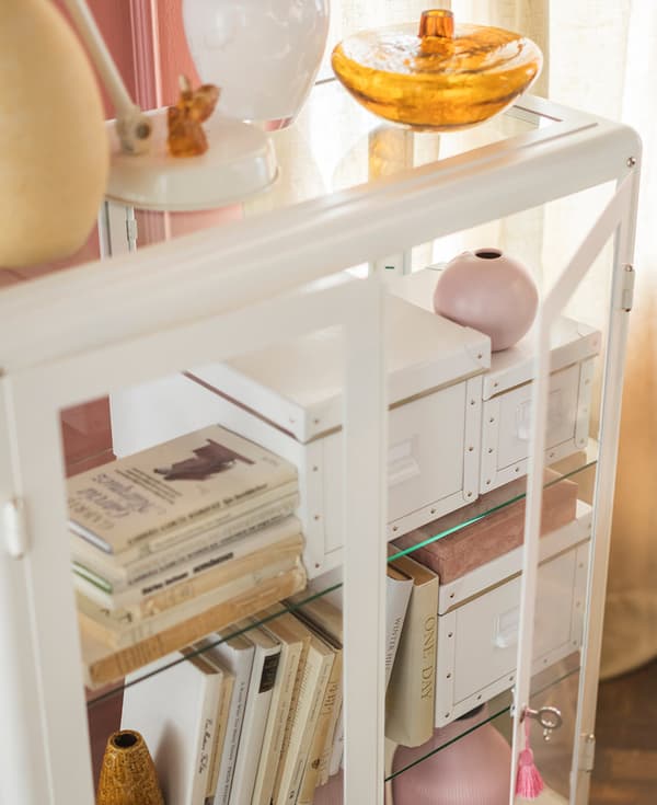 A white IKEA FABRIKÖR glass cabinet filled with books, boxes and vases.
