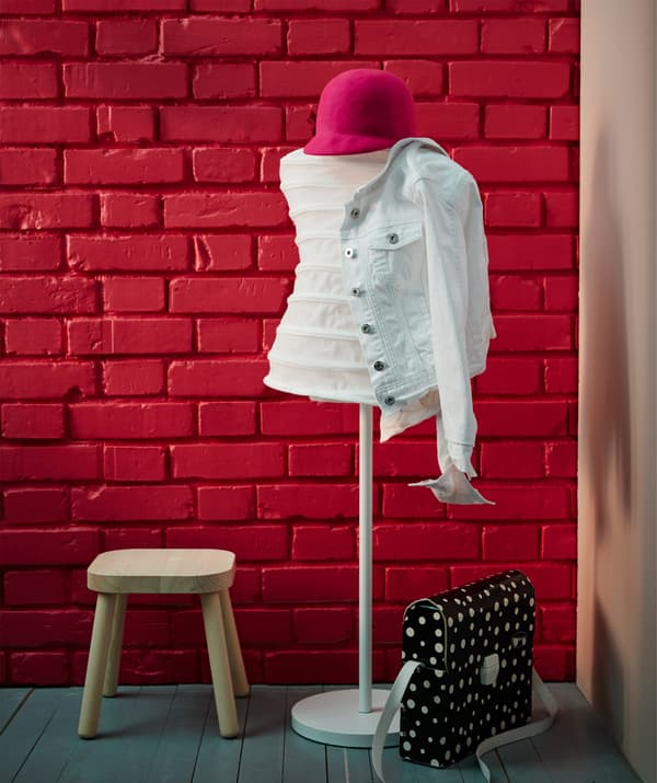 A white IKEA NÄPEN mannequin coat rack and stools in front of a red brick wall in a hallway.
