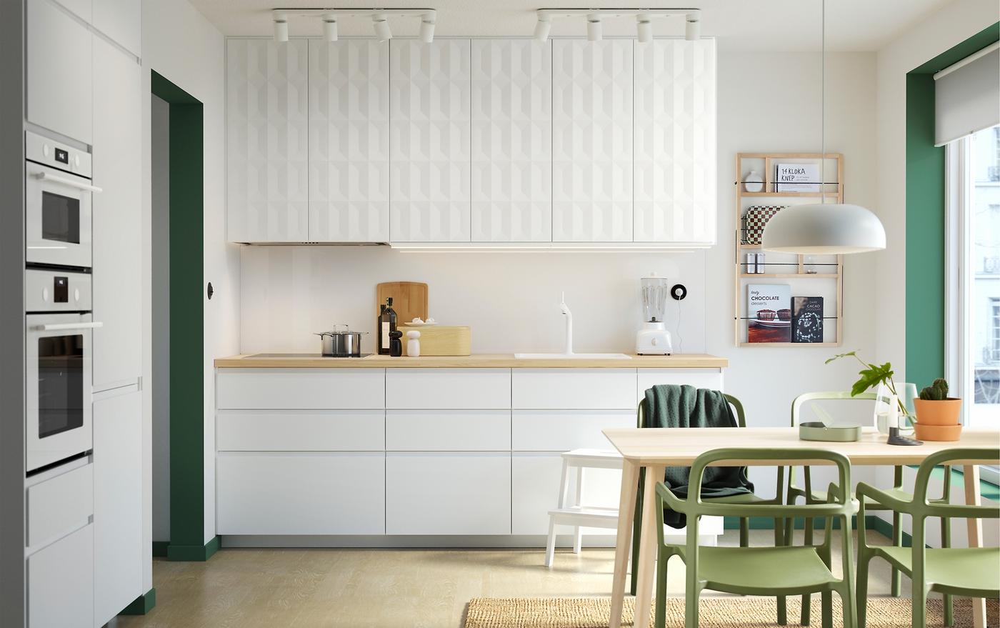 A white kitchen with green accents. Sleek white VOXTORP cabinet doors can be seen on the lower section and 3-dimensional HERRESTAD cabinet doors on the upper section. An LISABO table in ash veneer and YPPERLIG chairs in green can be seen in the foreground