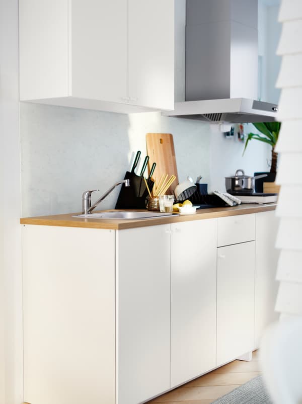 A white KNOXHULT kitchenette with a wooden worktop, potted herbs, pots, knives and various kitchen accessories.
