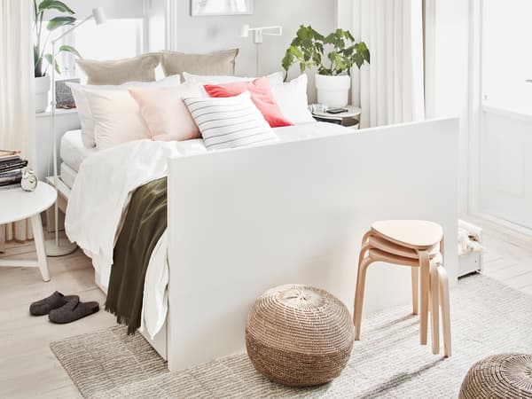 A white MALM double bed, made with many pillows and cushions and having its headboard as foot end, stands in a bright room.